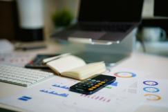 An illustrative photo of a workspace setup with a laptop, a calculator, an open notebook.