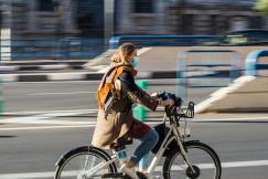 An illustrative photo of a woman in brown jacket wearing a mask and riding on bicycle on road 