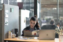 An illustrative photo of a woman reading a book while sitting at a desk with a laptop