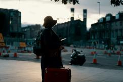 An illustrative photo of a woman wearing a coat standing on a road with travel luggage during the daytime