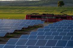 An illustrative photo of a field of solar panels in the foreground with a red electric train passing by in the background
