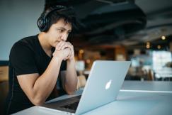 An illustrative photo of a man wearing headphones while sitting on chair in front of MacBook