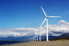 An illustrative photo of windmills standing in a row near the body of water with mountains in the background
