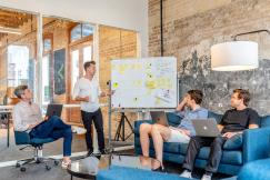 An illustrative photo of three men sitting with laptops and watching man beside whiteboard