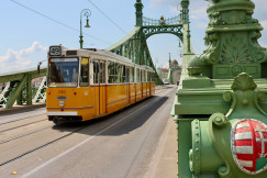 An illustrative photo of a yellow train traveling over a bridge in Budapest on a sunny day