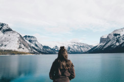 An illustrative photo of a woman facing a lake with mountains in the background.