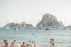 An illustrative photo of people on the beach with boats in the sea in the background.