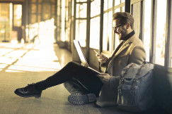 An illustrative photo of a man sitting near a window holding a phone and a laptop.