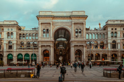 An illustrative photo of people walking near a big beige building with an arch.