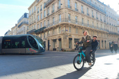 An illustrative photo of a couple of people riding a bike with a modern tram in the background.