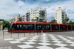 An illustrative photo of a red and black train traveling past tall buildings.