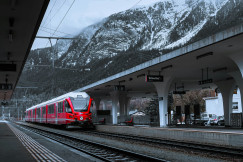 An illustrative photo of a red train traveling down train tracks next to a snow covered mountain.