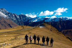 An illustrative photo of a group of people standing on a brown field near a snow covered mountain.