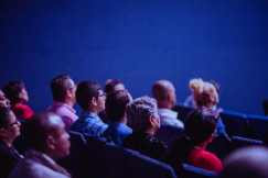 Audience seated and watching a presentation in a darkened conference or auditorium.