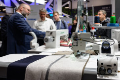 Industrial sewing machine stitching fabric at a trade fair, with attendees observing in the background.