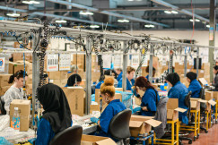 Female workers seated along an assembly line in a large factory next to cardboard boxes. They wear blue uniforms and sit at individual stations with tools and hanging cables overhead inside an industrial warehouse.