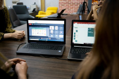 People sitting at a wooden table, listening to a webinar online meeting on two computer screens.