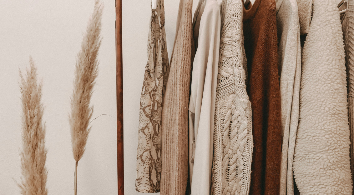 Neutral-toned sweaters, coats, and tops hanging on a clothing rack beside dried pampas grass.