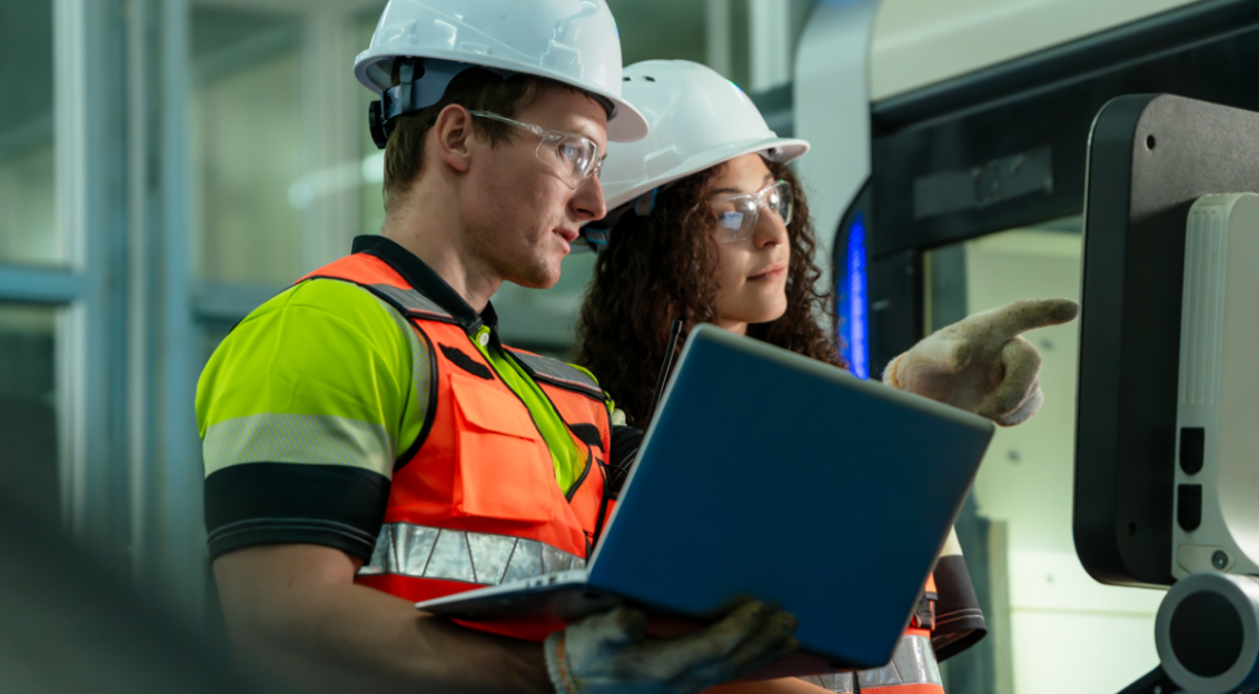 Two workers in safety gear inspect industrial equipment using a laptop.