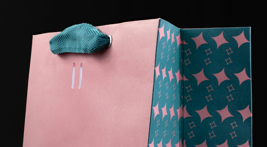 Pink and teal gift bag with a woven textile handle and decorative pattern against a black background.