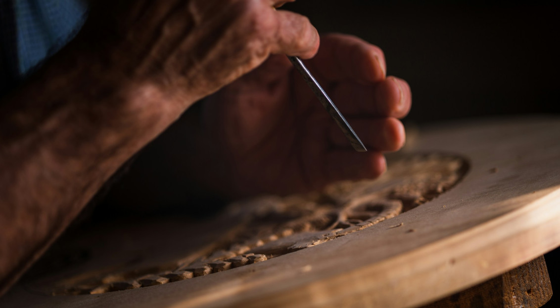 The photo presents a close-up of a person using a chisel to carve a decorative pattern into wood.