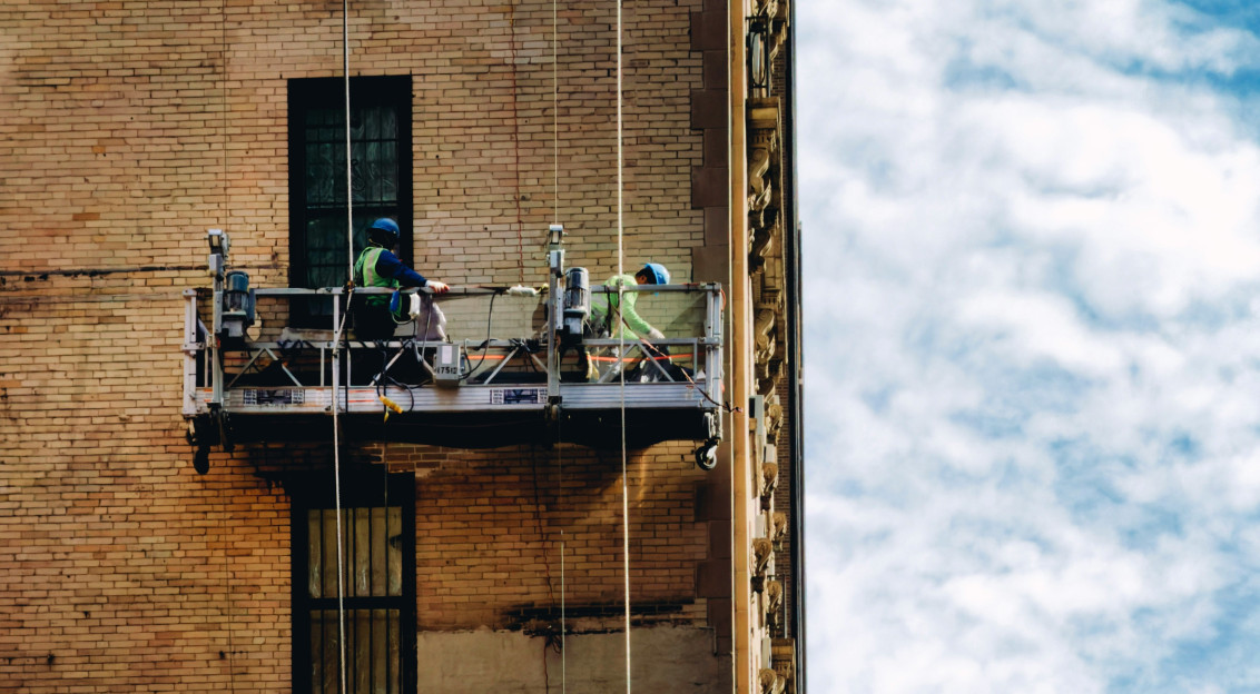 The picture shows the side of a tall brick building with two workers suspended on a motorized platform attached by vertical cables.