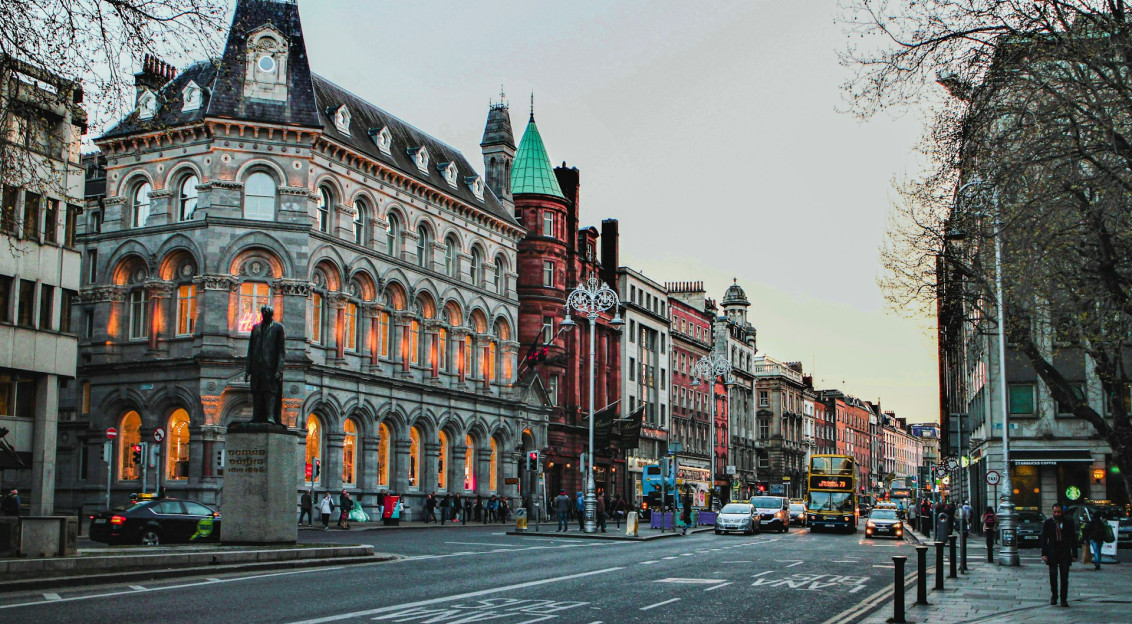The photo presents a Dublin street lined with historic buildings, with double-decker buses, cars, pedestrians and a statue in the foreground.