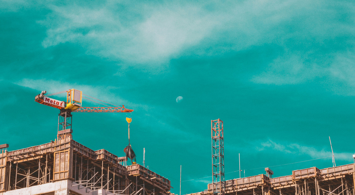 This picture shows a building under construction, featuring exposed concrete floors, scaffolding, and a tower crane lifting materials.