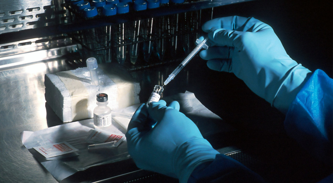 The photo presents blue-gloved hands holding a syringe and a small vial at a laboratory workstation, with racks of test tubes in the background.