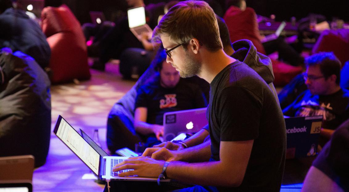An illustrative photo of a men using laptop in a room with many people