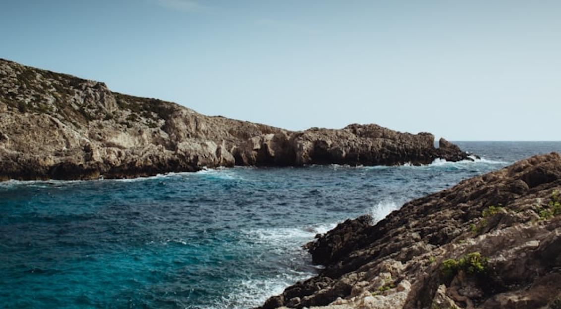 Photo by Unsplash An illustrative photo of a dramatic brown rock formation near the sea, with waves crashing against its base under a clear sky