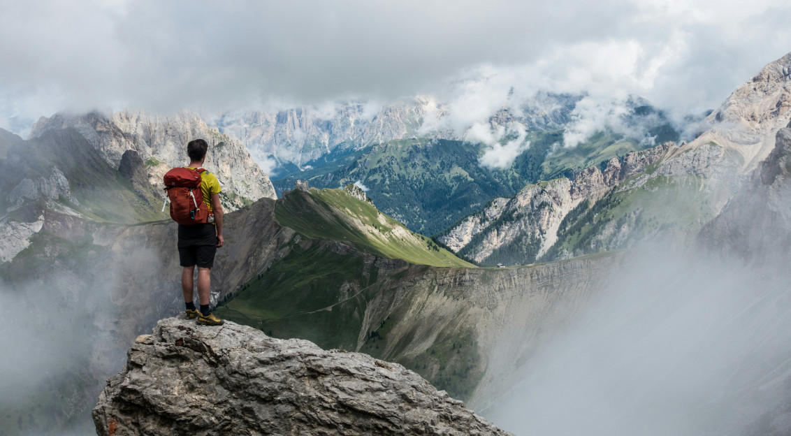 Photo by Unsplash An illustrative photo of a man with red backpack standing on cliff facing mountains