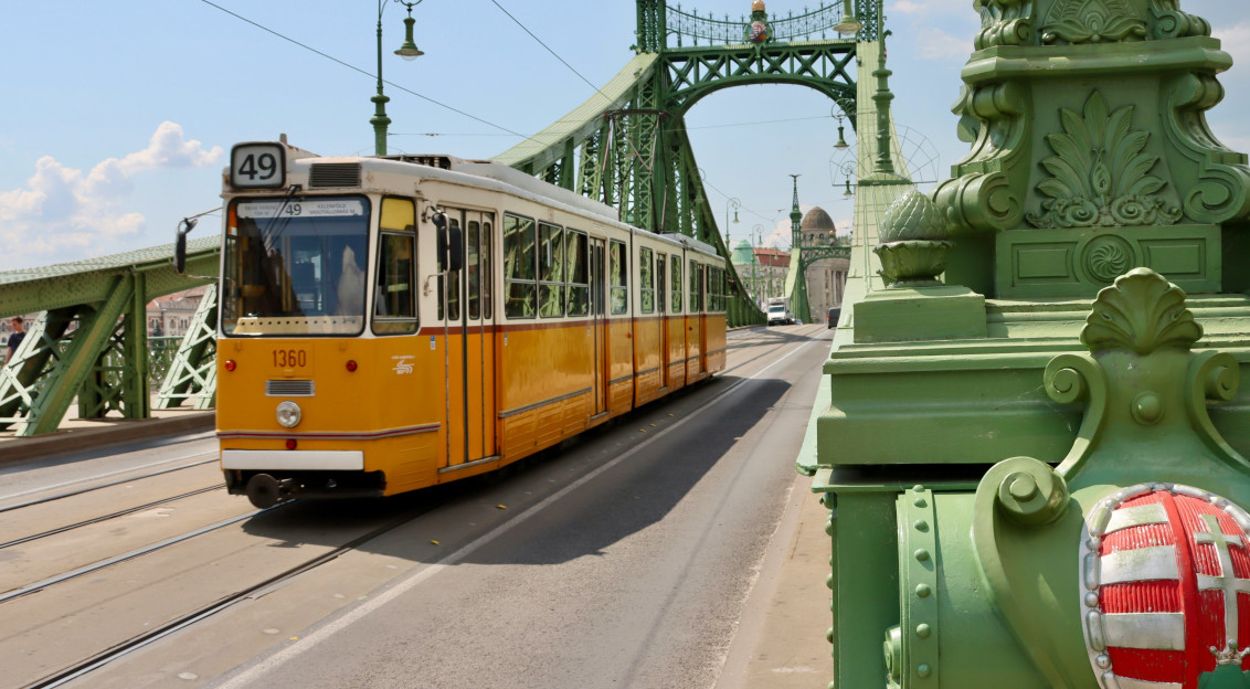 An illustrative photo of a yellow train traveling over a bridge in Budapest on a sunny day