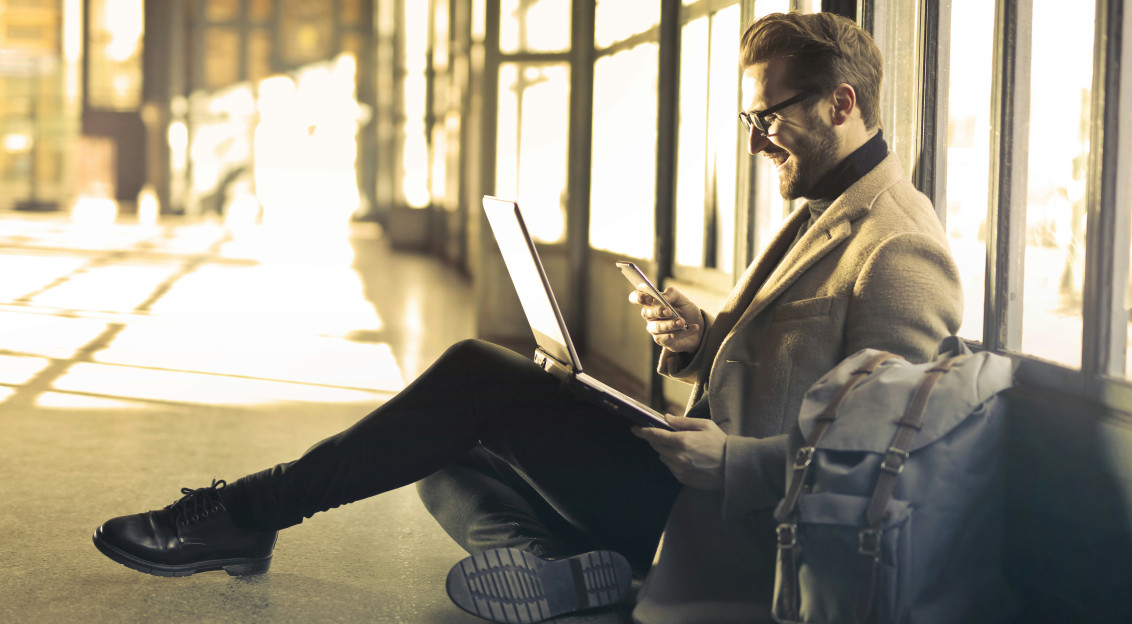 Image by Unsplash An illustrative photo of a man sitting near a window holding a phone and a laptop.