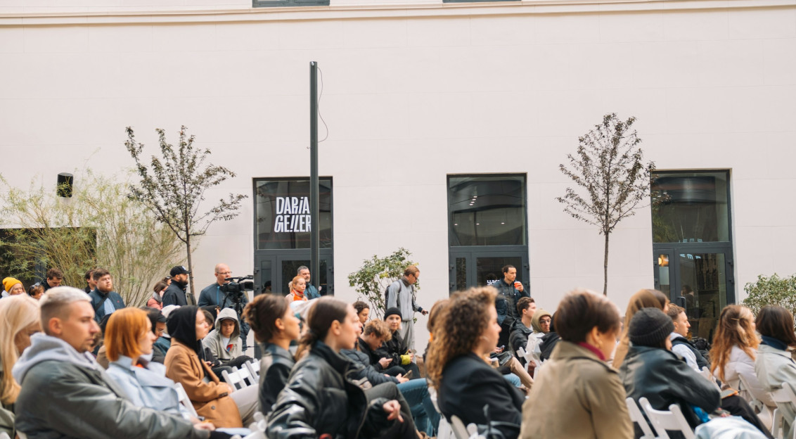 Audience seated outdoors at an event in a courtyard, listening attentively during a public talk or presentation.