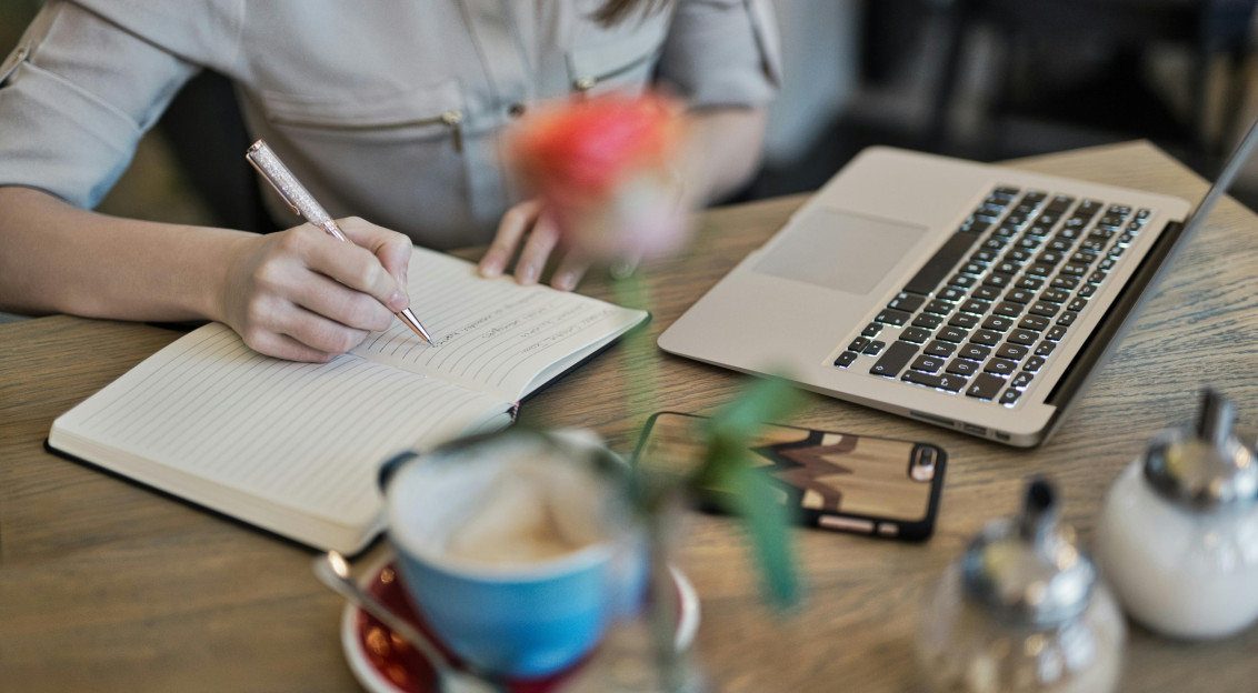 Person writing notes in a notebook beside a laptop and coffee, suggesting focused work or study in a café setting.