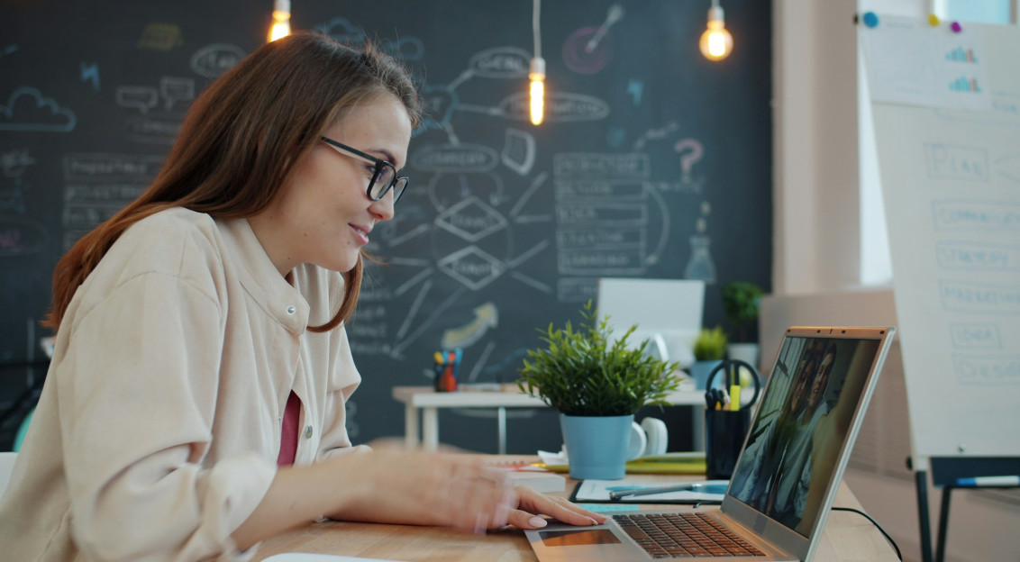 Woman working on a laptop at a desk, with a chalkboard full of diagrams in the background.