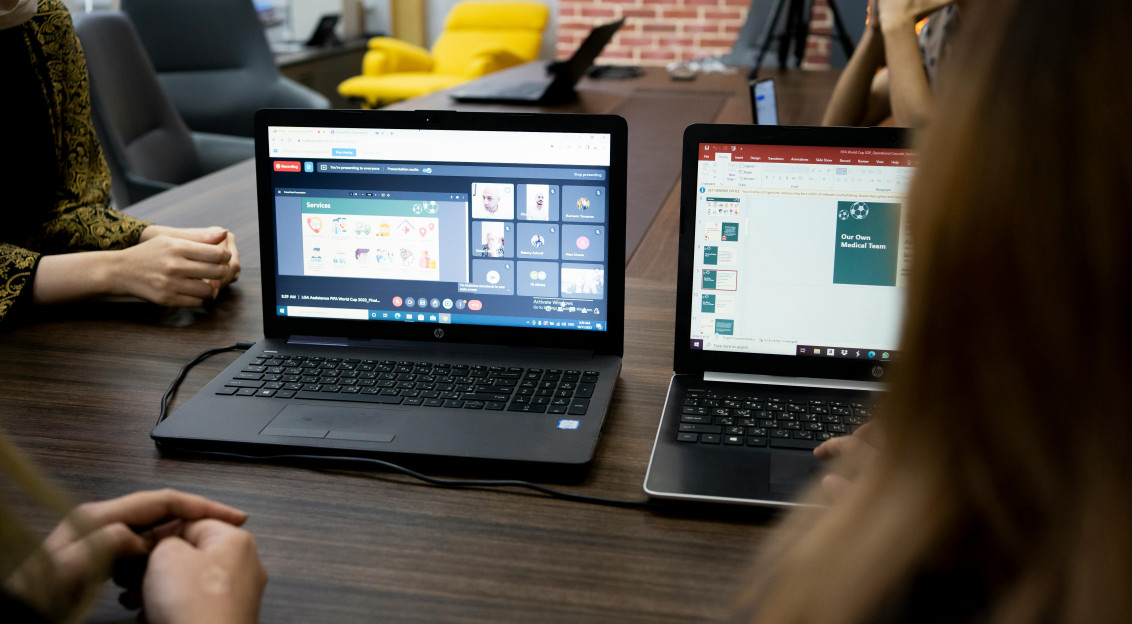 People sitting at a wooden table, listening to a webinar online meeting on two computer screens.